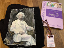 Folded Mark Twain T-shirt next to Tom Sawyer book, reading glasses and artist palette – literary tribute