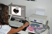 Artist seen from behind sitting at a computer editing T-shirt designs in Photoshop, with freshly printed images by the printer on the right
