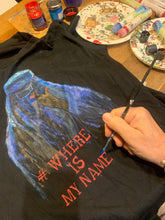  Artist’s hand painting the portrait of a veiled Afghan woman on a t-shirt, with paint jars and palette visible beside the artwork.