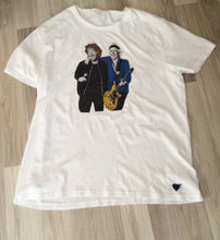 Full-body white hand-painted t-shirt showineg Mick Jagger holding a microphone with Keith Richards’ arm on his shoulder, wearing glittery black and electric blue outfits.