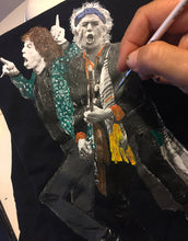 Artist’s hand painting a detailed portrait of Mick Jagger and Keith Richards on a white t-shirt, showcasing the hand-crafted, hand-painted artistic process.