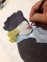 Artist’s hand painting the Tamara de Lempicka portrait on a t-shirt, with paint jars and palette visible.

