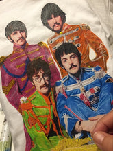 Artist’s hand painting The Beatles in Sgt. Pepper outfits on a t-shirt, with visible palette and color jars.
