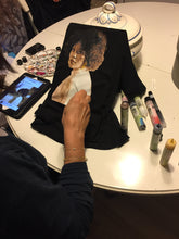 Artist seated at table painting a portrait of a black queen  customer on a T-shirt – close-up of arm and hand with brushes and fabric paints